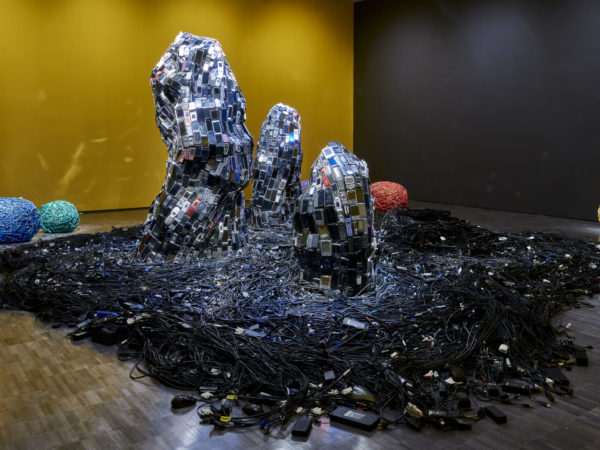 Three rock-like sculptures covered in old cell phones surrounded by a sea of cables and rocks made out of brightly colored ethernet cords.